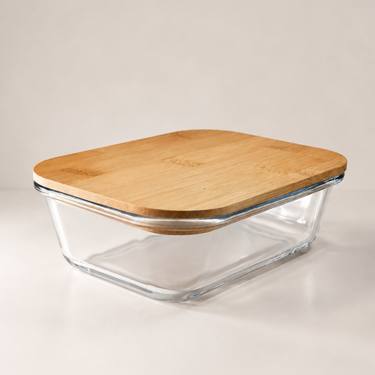 A clear glass storage container with a square shape and a light natural bamboo lid, resting on a white tabletop against a neutral beige background.